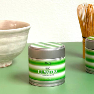 Matcha tin on a green table beside a ceramic tea bowl and a bamboo whisk for preparing matcha